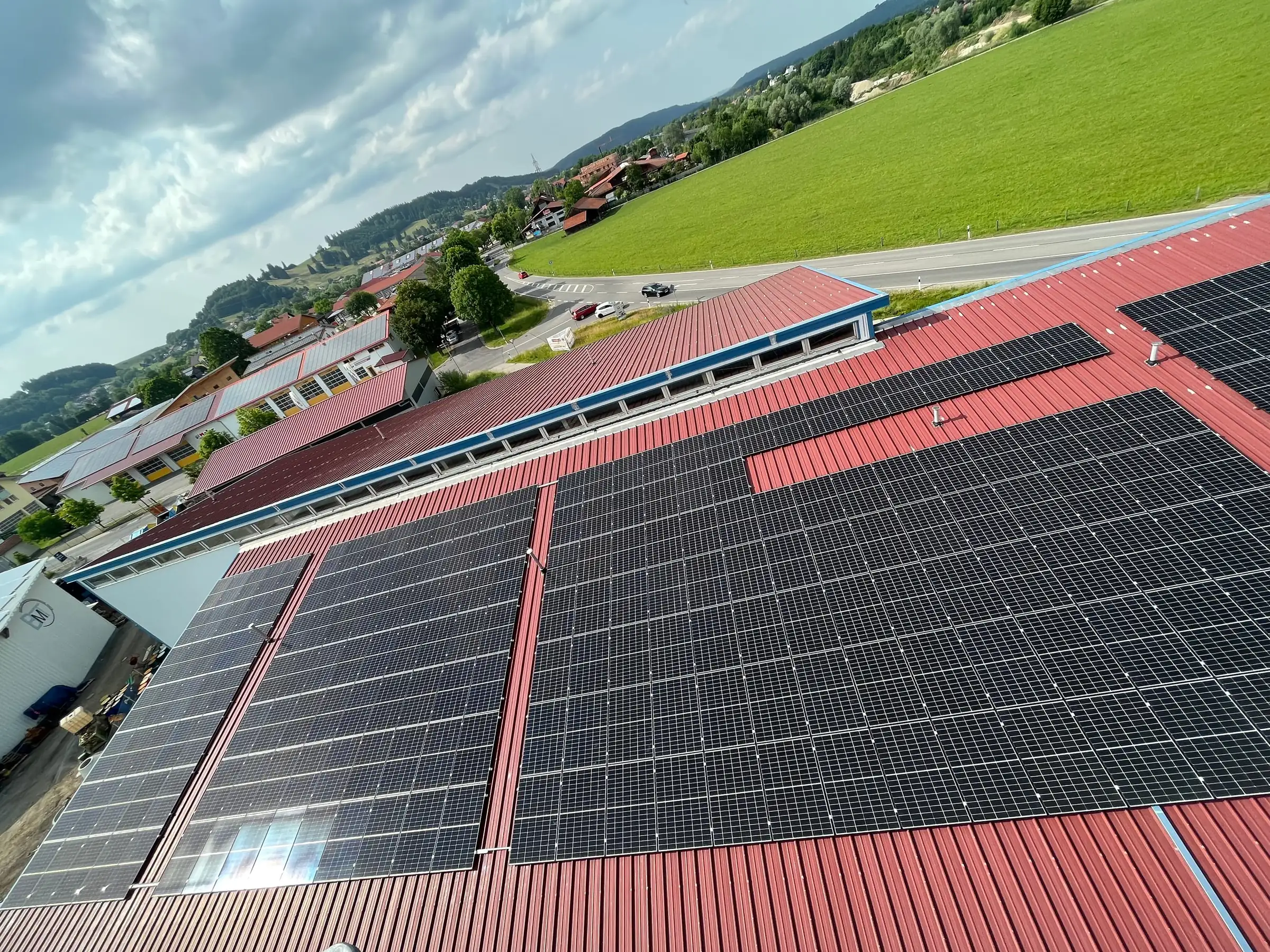 Großflächige Photovoltaikanlage auf den roten Trapezblechdächern eines Gewerbebetriebs, dahinter Wiesen und Hügel