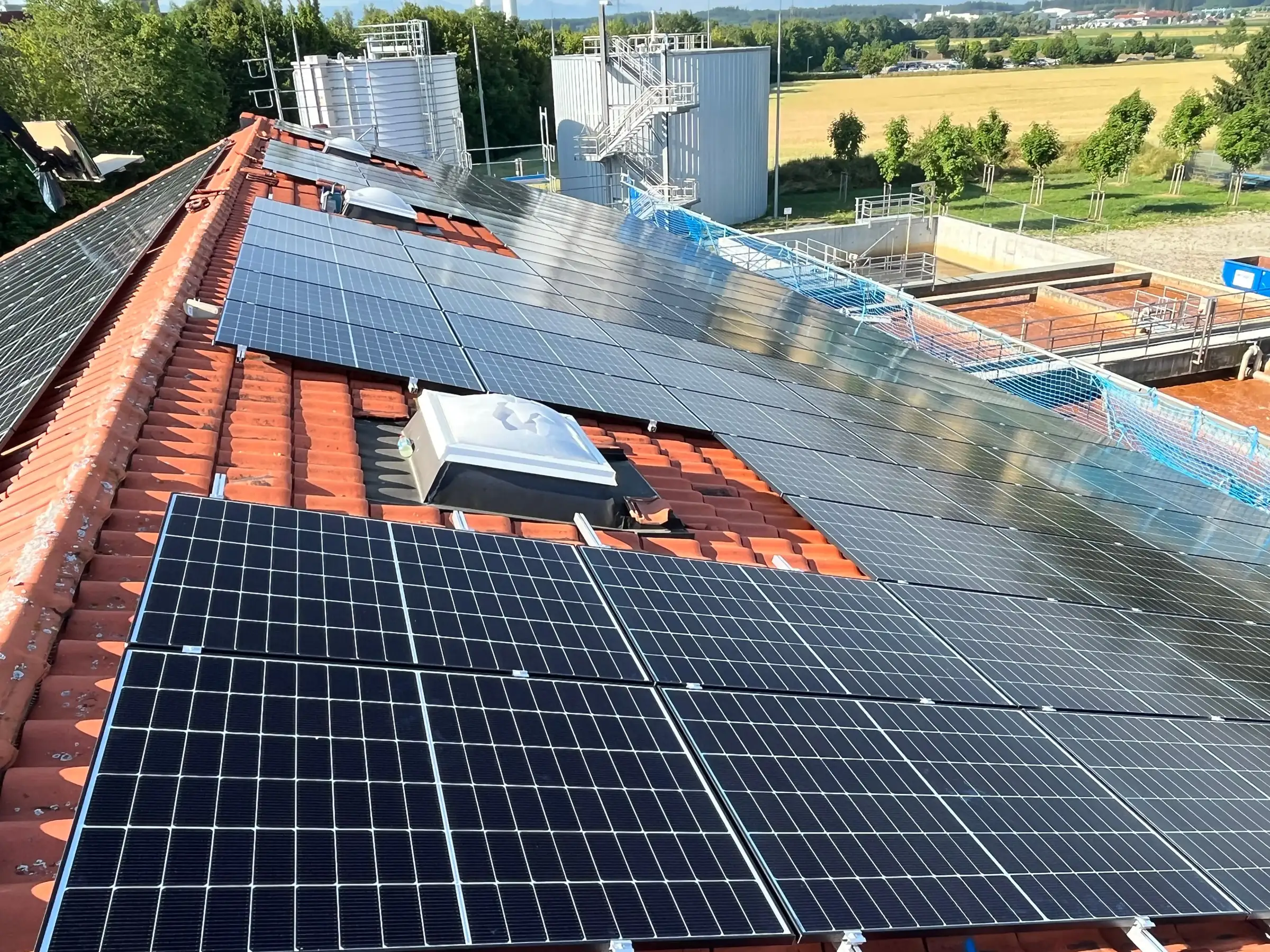 Photovoltaikmodule auf dem Ziegeldach eines Gebäudes der Kläranlage Hochland, dahinter Klärbecken und Behälter