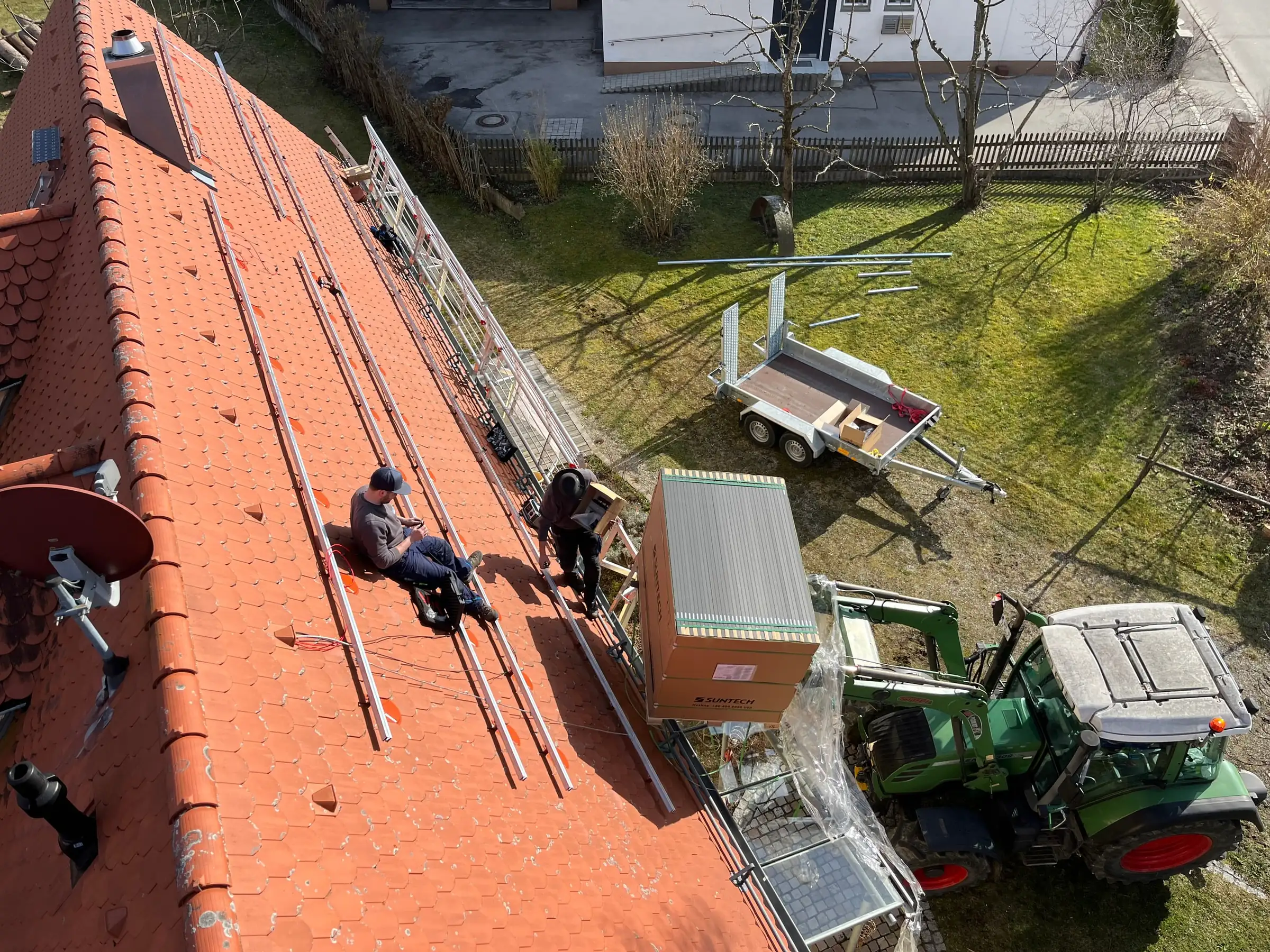 Luftaufnahme: Monteure setzen die Unterkonstruktion für eine Photovoltaikanlage auf ein Ziegeldach, unten Traktor mit Modulpaletten